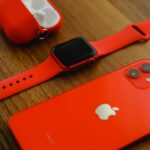 Close-up of red Apple products including iPhone, watch, and AirPods on a wooden surface.