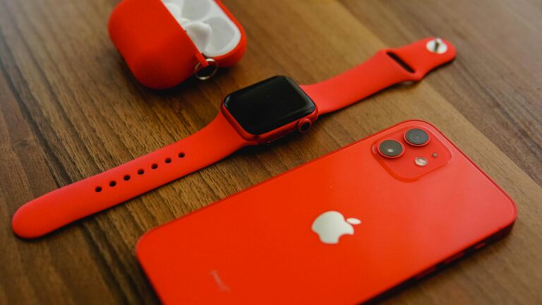 Close-up of red Apple products including iPhone, watch, and AirPods on a wooden surface.