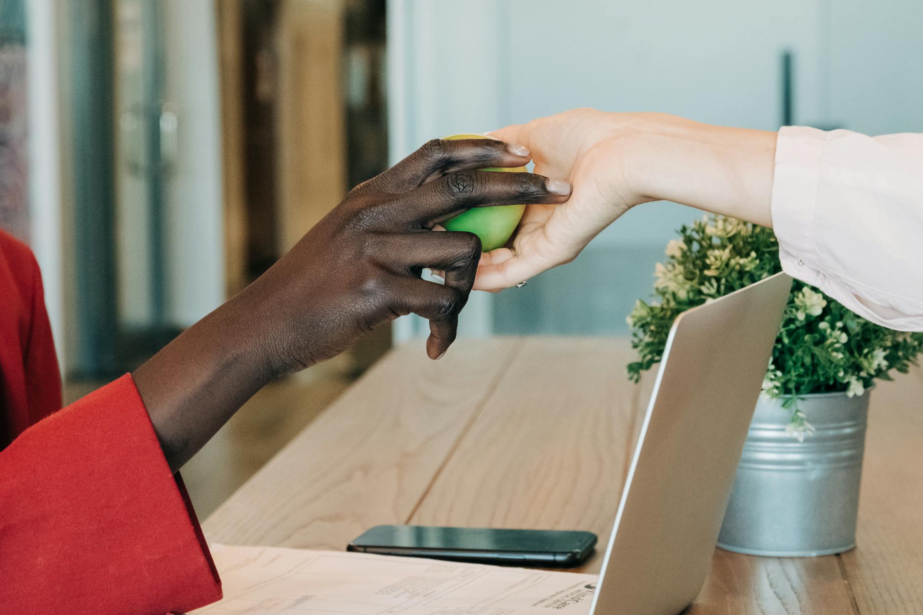 Close-up of hands exchanging an apple across a desk with a laptop and smartphone, symbolizing collaboration and healthy lifestyle at work.