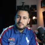 A young male vlogger in a home studio setup with a microphone and computer screen.
