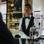 A bartender in formal attire serves a customer in a classic, stylish bar setting.