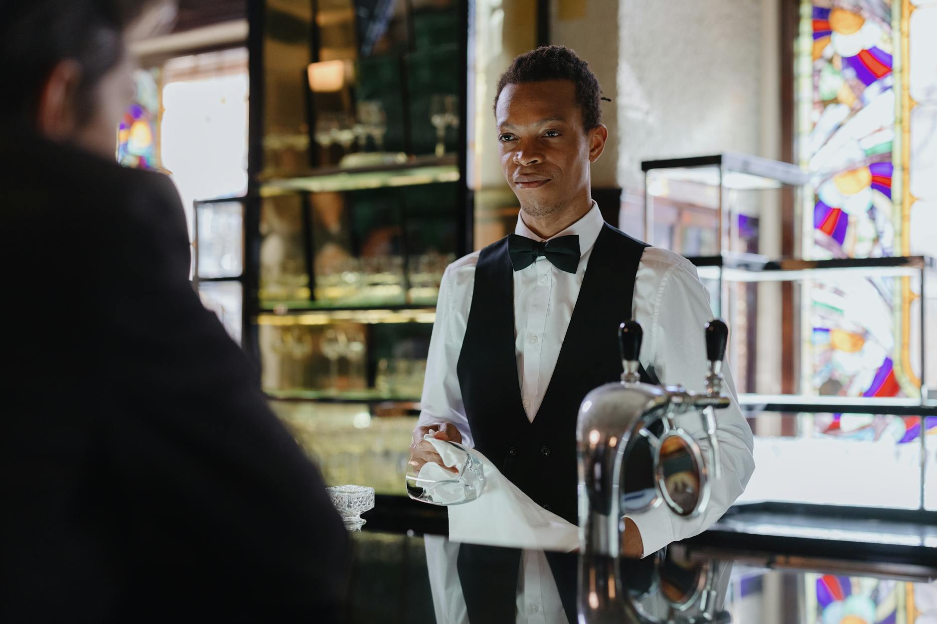A bartender in formal attire serves a customer in a classic, stylish bar setting.