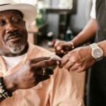 Senior man with a hat giving a tip to a young barber indoors, wearing bracelets and a watch.