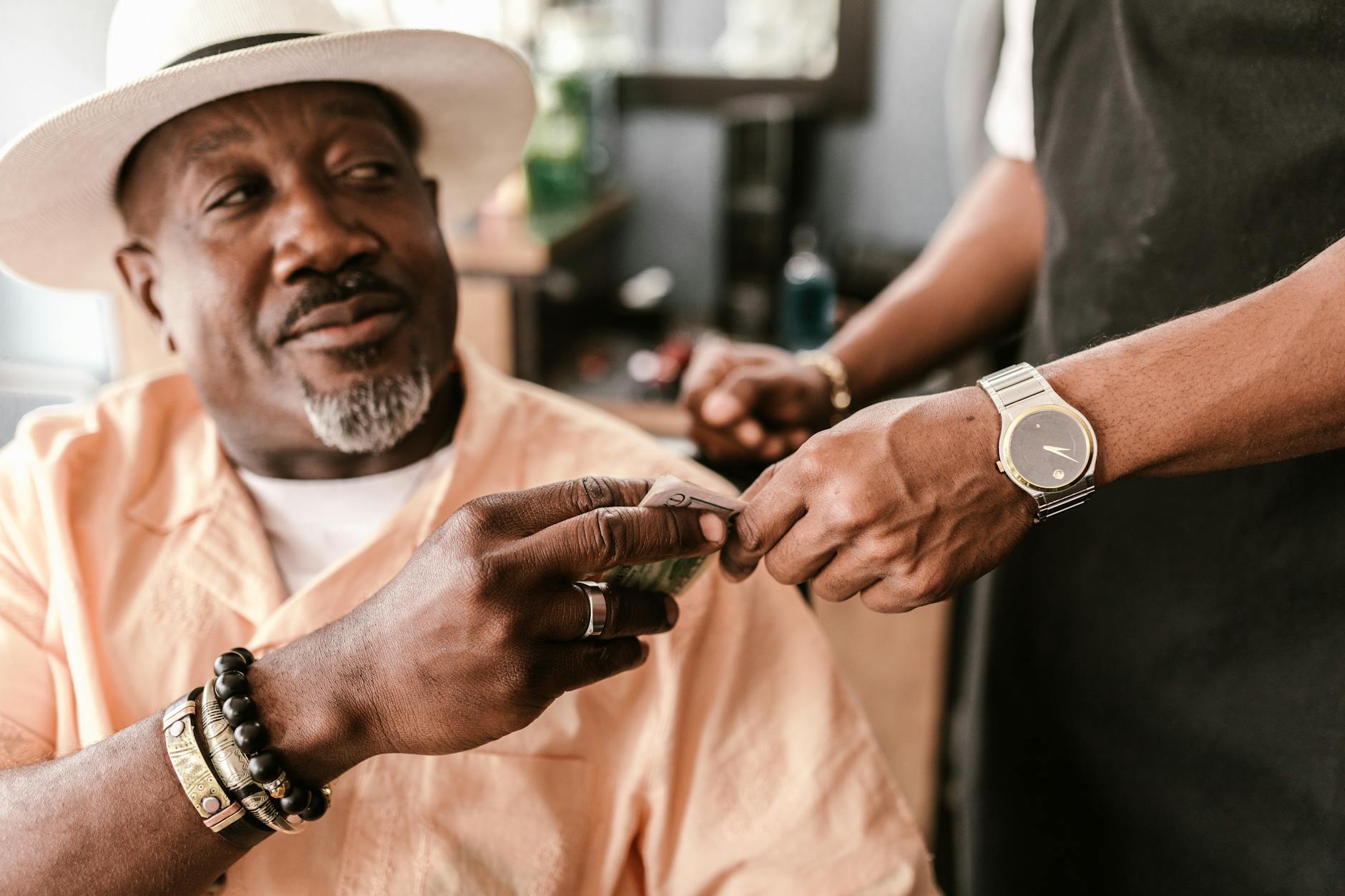 Senior man with a hat giving a tip to a young barber indoors, wearing bracelets and a watch.