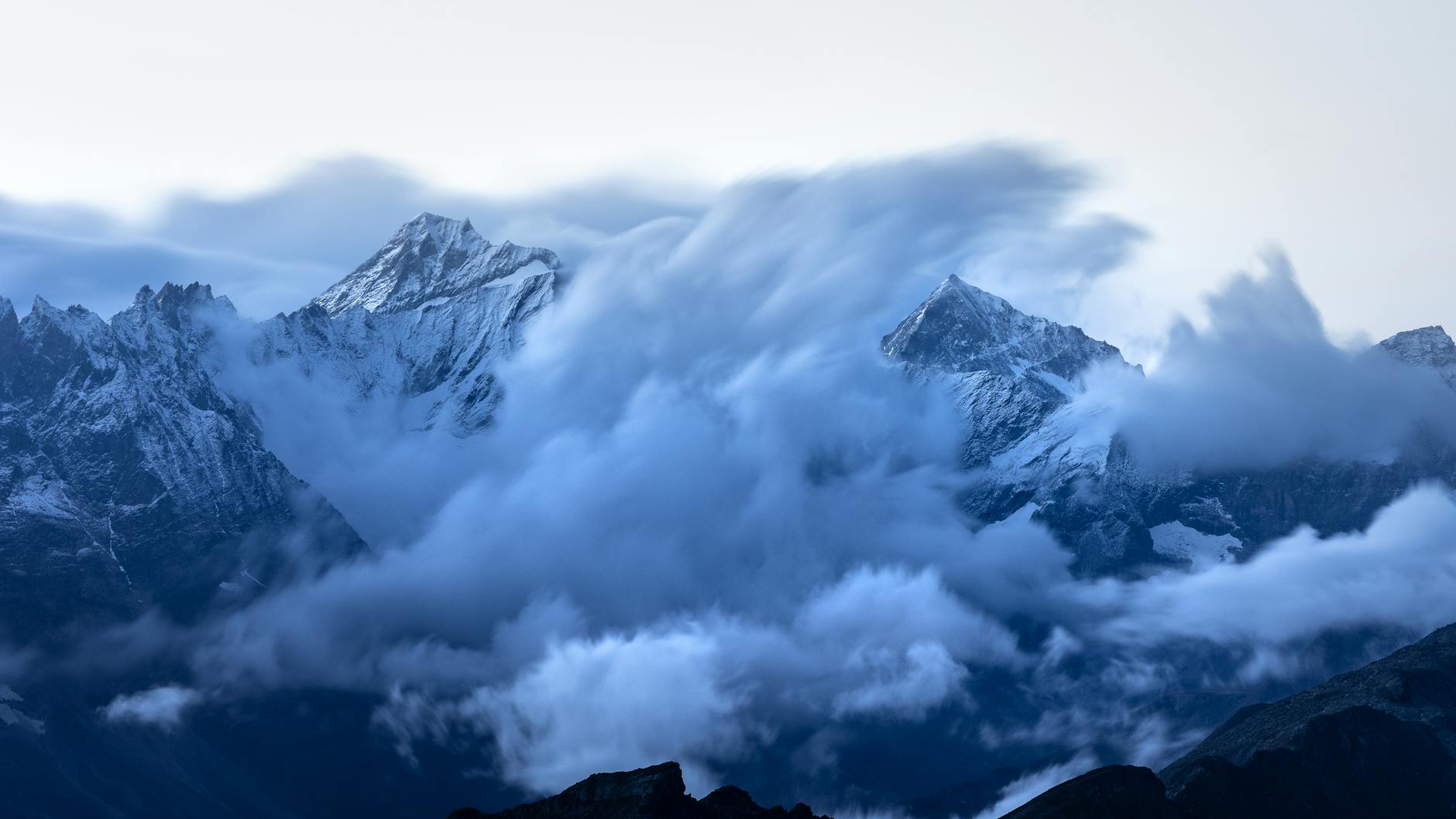 Stunning view of clouds swirling around snow-capped mountain peaks at twilight, creating a majestic landscape.